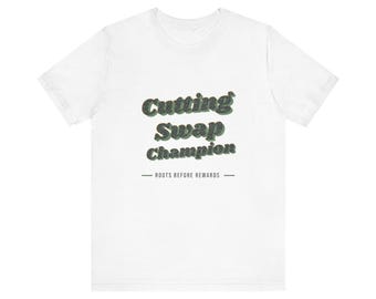 Può includere: Una t-shirt bianca a maniche corte con il testo Cutting Swap Champion in un carattere testurizzato verde scuro. Sotto il testo principale, la frase ROOTS BEFORE REWARDS è stampata in un carattere nero semplice con linee orizzontali.