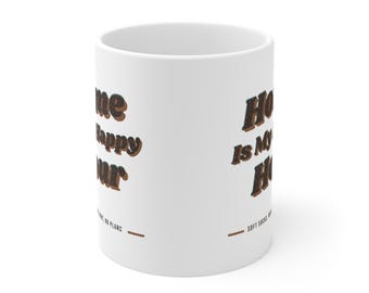 May include: A white ceramic mug featuring the text Home is my happy hour in a dark brown, textured font. Below the main text, smaller black lettering reads soft socks, wine, no plans, accompanied by two horizontal brown lines on either side.