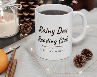 May include: A white ceramic mug featuring the dark brown text Rainy Day Reading Club, a horizontal line, and the smaller text TEA REFILLED, PHONE OFF with a small blue star. The mug has a standard handle and is filled with a dark liquid.