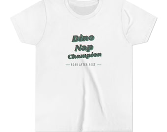 May include: A white short-sleeved t-shirt featuring the text "Dino Nap Champion" in a dark green, textured font with a subtle red outline, followed by the text "— ROAR AFTER REST —" in a smaller, simple dark green sans-serif typeface.