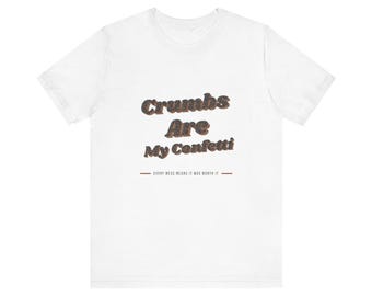 以下が含まれることがあります： 暗い質感のフォントで「Crumbs Are My Confetti」というテキストがプリントされた白い半袖Tシャツ。その下には、茶色の水平線で囲まれた「EVERY MESS MEANS IT WAS WORTH IT」という小さなテキストが配置されています。