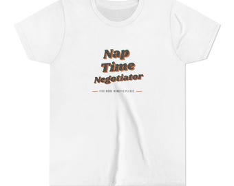 Può includere: Una t-shirt bianca a maniche corte con il testo Nap Time Negotiator in un carattere scuro e testurizzato con contorni arancioni. Sotto il testo principale, una riga più piccola riporta FIVE MORE MINUTES PLEASE tra due linee orizzontali arancioni.