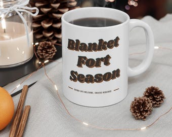 May include: A white ceramic mug featuring the text Blanket Fort Season in a dark brown, textured font. Below the main text, smaller lettering reads GROWN-UPS WELCOME, SNACKS REQUIRED, flanked by two horizontal lines. The mug has a standard handle.