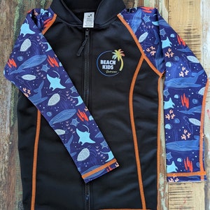 May include: Black long-sleeve rash guard with orange trim and a colorful "Beach Kids Swimwear" logo. The rash guard features a blue and white print of sea creatures, including turtles, stingrays, and starfish.