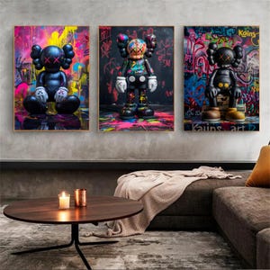 Peut inclure: Trois impressions d'art encadrées représentant des personnages stylisés avec des yeux en forme de X sur des fonds de graffiti. Les personnages sont noirs avec des accents colorés, et les graffitis incluent des éclaboussures de rose, de jaune et de bleu.
