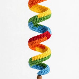 May include: A hanging spiral decoration crafted from crocheted yarn in a repeating rainbow color sequence of red, orange, yellow, green, and blue. The spiral features a small wooden bead at the bottom, finished with a multi-colored yarn tassel.