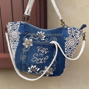 May include: A blue denim tote bag with a white crocheted trim and a white crocheted strap. The bag has a pocket on the front with the text "Kiss My Soul" embroidered in gold thread. The bag is decorated with white beads and white flowers.