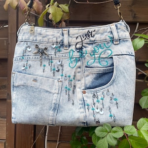 May include: A light blue denim bag with a black strap and turquoise embroidery. The bag has a large pocket on the front with the words "Just be you" embroidered in black and turquoise thread.