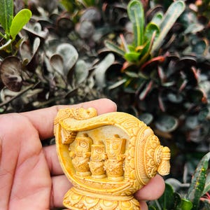 Puede incluir: Una detallada figura tallada en tonos amarillos que presenta tres figuras dentro de una estructura ornamentada en forma de barco. La talla muestra intrincados patrones y diseños, sobre un fondo de follaje verde. La figura se sostiene en una mano.