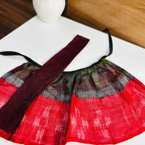 May include: A flared skirt featuring a vibrant red and black ikat pattern with a textured weave. The garment includes a solid dark maroon waistband with attached black fabric ties on each side, designed for a wrap-around fit with a gathered silhouette.