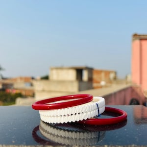 Puede incluir: Tres pulseras apiladas sobre una superficie reflectante. Dos son de un rojo vibrante y una blanca con borde festoneado. El fondo muestra un paisaje urbano bajo un cielo azul claro, con edificios y tejados visibles.