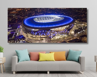 May include: A panoramic image of a modern stadium at night, lit with blue and white lights. The stadium has a circular design and a prominent roof. The surrounding cityscape is visible with city lights.