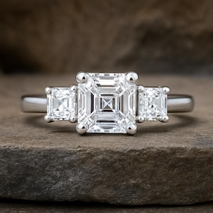 May include: A silver engagement ring with three square-cut diamonds. The central diamond is larger than the two side stones. The ring is set on a silver band, and the diamonds are clear and sparkling.