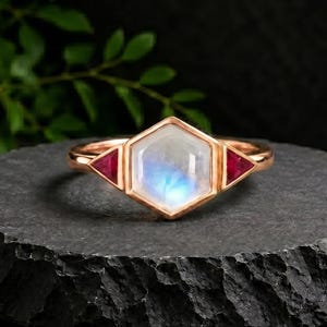 May include: A rose gold ring featuring a central hexagonal moonstone cabochon flanked by two small triangular red rubies. The band is polished metal, and the setting holds the stones in a geometric arrangement with a smooth, reflective finish.