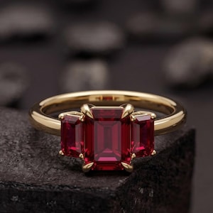 Op de afbeelding: Een gouden ring met drie rechthoekige, dieprode edelstenen. De band en zetting van de ring zijn helder goudkleurig, wat een opvallend contrast vormt met de donkerrode stenen. De ring wordt getoond op een donker, getextureerd oppervlak.