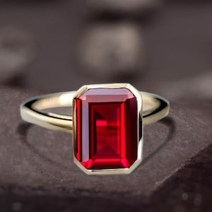 May include: A gold ring with a large, rectangular, faceted red gemstone. The gemstone is set in a bezel setting, and the band is a simple, polished gold. The ring is displayed on a dark, textured surface.