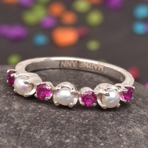 Könnte beinhalten: Ein silberner Ring mit Perlen und rosa Edelsteinen. Der Ring liegt auf einem dunkelgrauen Stein, mit bunten Konfetti im Hintergrund. Das Wort "MARNEANN" ist auf dem Ring eingraviert.