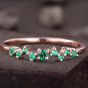 Puede incluir: Un delicado anillo de oro rosa con un diseño curvo que presenta una fila de pequeñas piedras preciosas de color verde esmeralda.