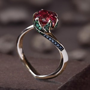 Puede incluir: Un anillo de oro con una piedra preciosa roja rodeada de piedras preciosas verdes. La banda está engastada con diamantes negros.