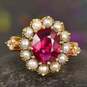 May include: A gold ring with a large oval ruby surrounded by a halo of pearls. The ring is set on a dark gray stone.