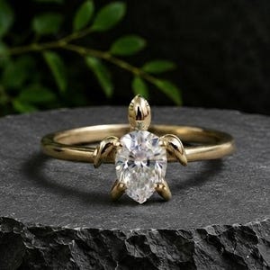 Op de afbeelding: Een gouden ring met een centrale peervormige, heldere edelsteen in een schildpad-vormige metalen zetting. Het schildpadontwerp heeft vier kleine pootjes en een kop, waarbij de peervormige steen het schild van de schildpad vormt.