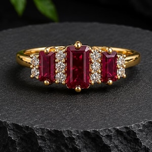 May include: A gold ring featuring three rectangular red rubies arranged horizontally, with small round white diamonds set between and around the rubies. The central ruby is larger than the two flanking rubies, all set in a polished gold band.