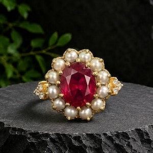 May include: A gold ring featuring a large, oval-cut deep red gemstone at the center, surrounded by a halo of small, round white pearls. The band is accented with tiny clear stones on each side, creating a detailed and ornate appearance.