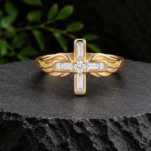 Puede incluir: Un anillo de oro con una pieza central en forma de cruz adornada con piedras transparentes de corte baguette y redondo. La banda del anillo está diseñada con motivos estilizados en forma de alas que se extienden desde la cruz central.
