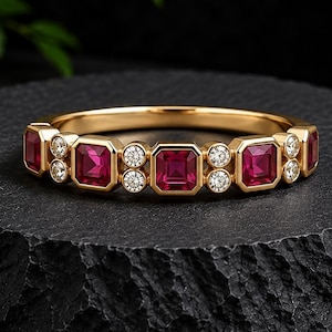 Pode incluir: Um anel de banda em ouro amarelo polido com um padrão repetitivo de rubis vermelhos de corte quadrado e pequenos diamantes brancos redondos de corte brilhante. As pedras preciosas estão cravadas em bisel ao longo da parte superior.