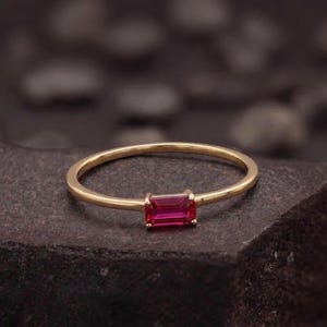 Puede incluir: Un anillo de oro con una gema rectangular de color rojo intenso. La banda del anillo es delgada y lisa, y la gema está engastada en un diseño sencillo y elegante. El anillo se muestra sobre una superficie oscura y texturizada.