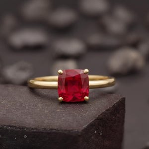 Op de afbeelding: Een gouden ring met een grote, vierkant geslepen, helderrode edelsteen. De edelsteen wordt op zijn plaats gehouden door vier kleine gouden pootjes. De ringband is van eenvoudig, gepolijst goud.