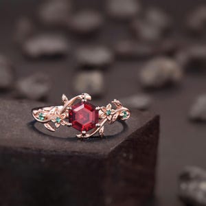 Könnte beinhalten: Ein Ring aus Roségold mit einem zentralen, sechseckigen roten Edelstein. Die Ringschiene ist mit detaillierten Metallmotiven aus Ranken und Blättern gestaltet, die durch kleine grüne Steine in den floralen Elementen akzentuiert werden.