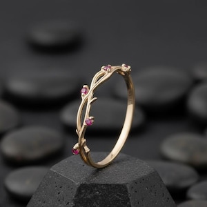 May include: A gold-colored ring with a delicate vine design. The ring features small, round, red gemstones set along the vine. The ring is displayed on a dark, textured surface, showcasing its intricate details and elegant style.