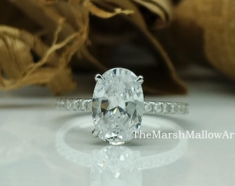 May include: A silver ring with an oval-shaped diamond and a band of small diamonds. The ring is on a white surface with a brown background.