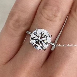 Puede incluir: Primer plano de una mano con un anillo de plata con un gran diamante redondo y transparente. El anillo tiene un engaste de pavé con diamantes más pequeños alrededor de la piedra central. La mano lleva esmalte de uñas rojo.