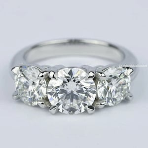 Puede incluir: Un anillo de compromiso de plata de tres piedras con un gran diamante central y dos diamantes más pequeños a cada lado.