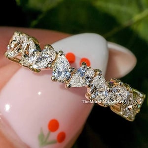 Könnte beinhalten: Ein Goldring mit einer Reihe von birnenförmigen, klaren Steinen. Der Ring befindet sich an einem Finger mit rosa Nagellack und einem Kirschblütenmuster.