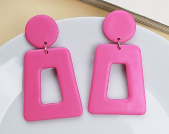 Puede incluir: Un par de pendientes geométricos de color rosa con forma cuadrada y parte superior redonda. Los pendientes están hechos de un material liso y mate.