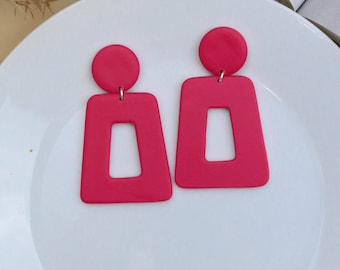 Puede incluir: Un par de pendientes llamativos de color rosa brillante con una forma geométrica rectangular y una parte superior redonda. Los pendientes tienen un recorte en el centro del rectángulo.