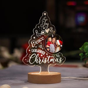 Może przedstawiać: Przezroczysta akrylowa lampka w kształcie choinki z drewnianą podstawą. Wzór zawiera napisy Joy, Love, Peace, Believe oraz Christmas w białym kolorze, a także gwiazdki i okrągłe zdjęcie przedstawiające trzy osoby.