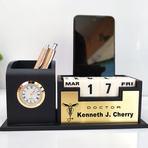 Puede incluir: Organizador de escritorio negro con un calendario dorado, un reloj y un portaplumas. El calendario muestra "MAR 17 FRI" y la placa dorada dice "DOCTOR Kenneth J. Cherry".