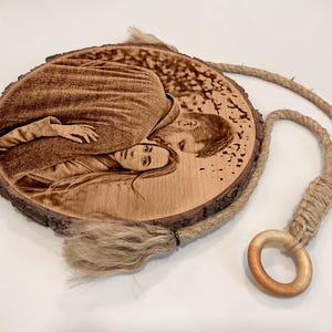 Puede incluir: Un disco de madera circular con un retrato grabado a láser de dos personas abrazadas. Se conservan los bordes de corteza natural. Una cuerda de yute gruesa de color tostado está sujeta a la parte superior y termina en un anillo de madera.