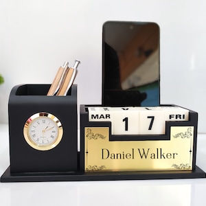 May include: Black desktop organiser with a clock, pen holder, calendar, and a mobile phone stand. The calendar displays "MAR 17 FRI" and the name "Daniel Walker" is engraved on the gold plate.