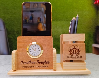May include: A wooden desk organizer with a clock, phone holder, and pen holder. The clock has a gold bezel and Roman numerals. The organizer is engraved with a name and title, and a company logo. A smartphone is in the phone holder.