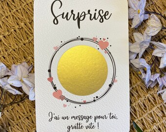 Pode incluir: Um cartão retangular branco de raspar com a palavra Surprise em letra cursiva preta no topo. Uma área circular dourada para raspar está rodeada por uma moldura circular preta com pequenos corações cor-de-rosa. O texto J'ai un message pour toi, gratte vite ! está impresso a preto na parte inferior.