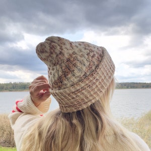 May include: A woman wearing a white and brown knitted beanie with a striped sweater. The beanie has a textured pattern and is pulled down over her ears. She is standing in front of a body of water with trees in the background.