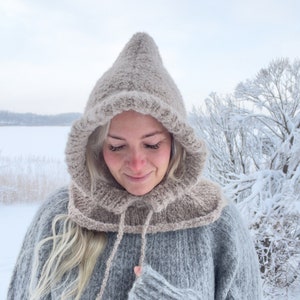 Könnte beinhalten: Ein hellbrauner Kapuzen-Kragen mit einem langen, fließenden Schal. Der Kragen ist aus weichem, flauschigem Garn gefertigt und hat ein schlichtes, elegantes Design. Der Schal ist um den Hals gebunden und hängt an der Vorderseite des Körpers herunter.