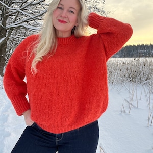 Może przedstawiać: Kobieta w jasnym pomarańczowym, puszystym swetrze i ciemnoniebieskich dżinsach. Stoi w śnieżnym krajobrazie.