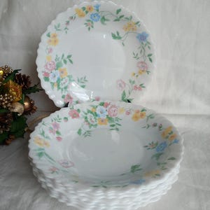 Puede incluir: Una pila de cuencos blancos con bordes festoneados y un diseño floral. Los cuencos presentan un delicado patrón de flores rosas, amarillas y azules con hojas verdes. Los cuencos están apilados sobre una superficie blanca.