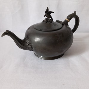 Peut inclure: Une théière en étain vintage avec un bouton en forme d'oiseau sur le couvercle. La théière a une patine usée et vieillie et un corps légèrement arrondi.